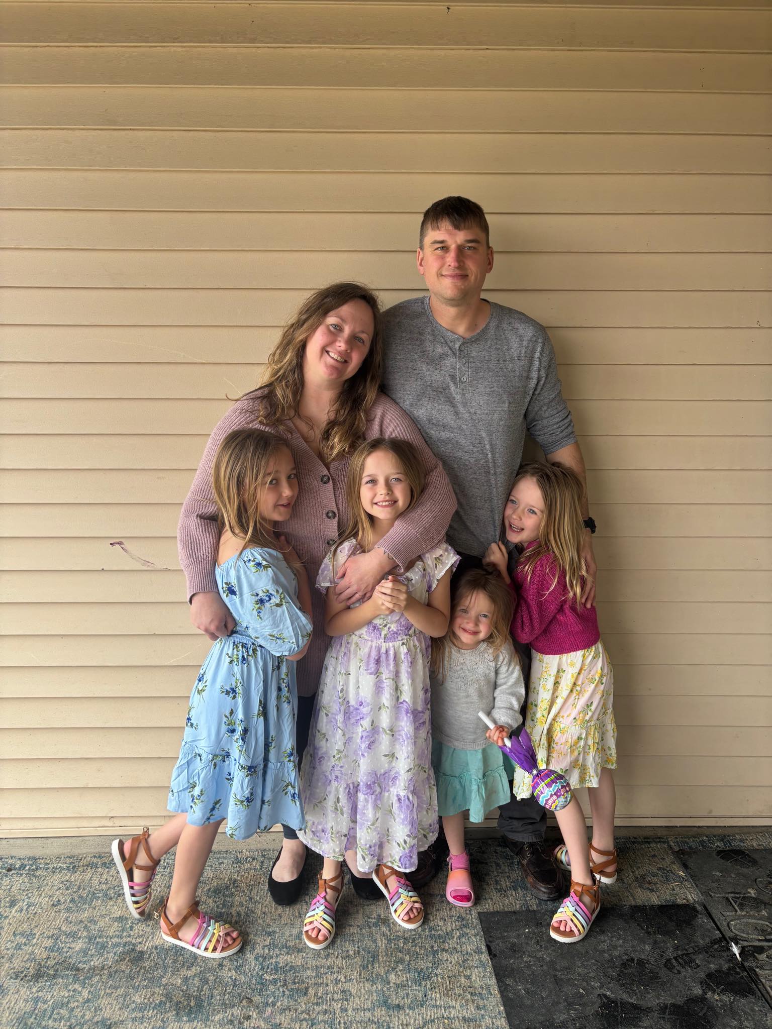 644267091_10229151714208756_8550147004314958734_n Family portrait featuring a couple and their four young daughters, all smiling and dressed in colorful spring dresses. The group is posed against a neutral wall, showcasing a warm family bond and joyful atmosphere.